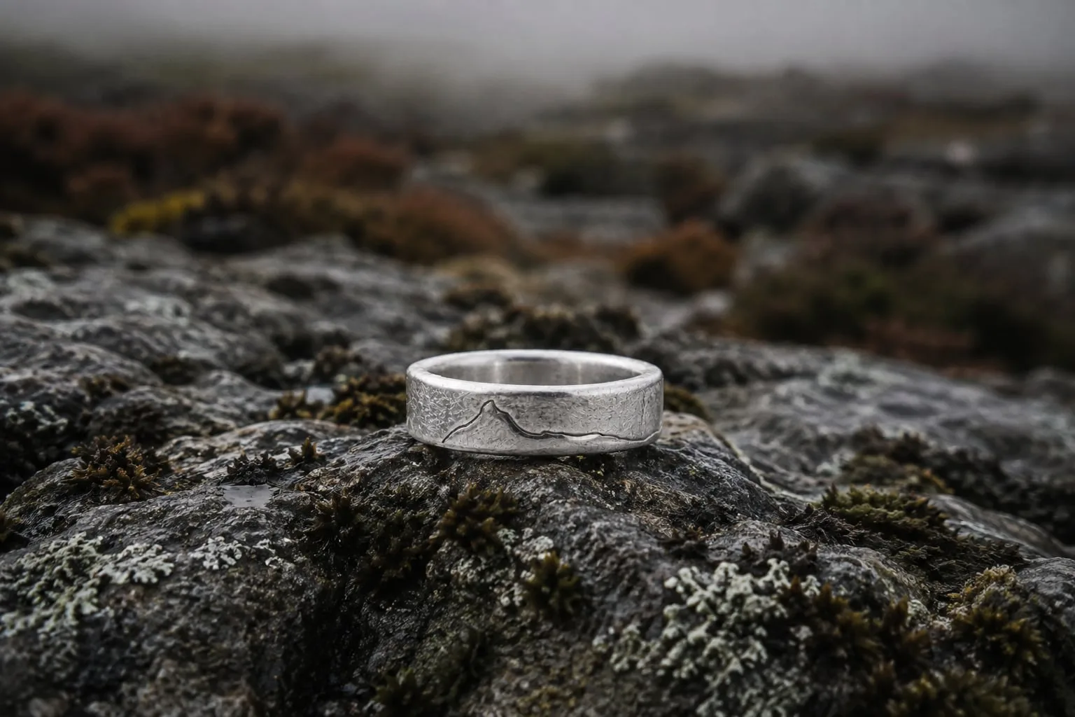 The Yorkshire Three Peaks sterling silver ring, its fine engraved ridgeline visible, resting on wet lichen-covered stone.