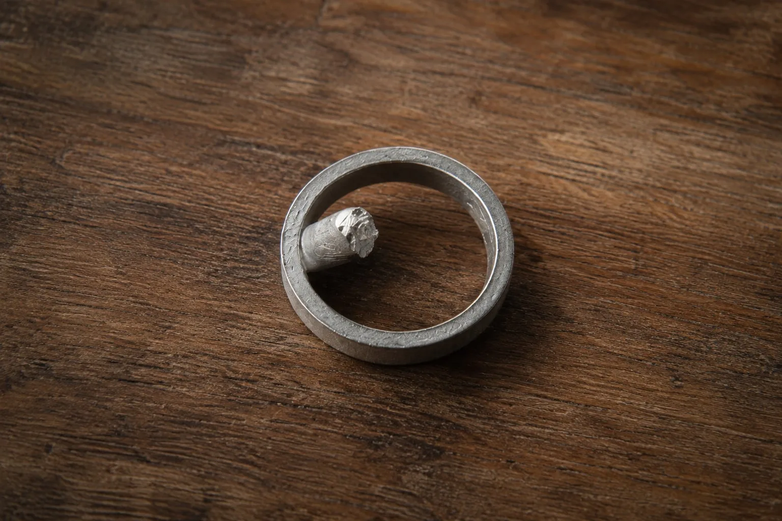 A rough silver ring cast, still attached to its sprue, resting on a wooden bench, its surface textured from the investment mould.